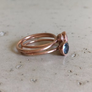May include: Three copper rings stacked together. Two rings have bezel-set gemstones: one pink and one teal. The rings are on a light-colored, textured surface. The rings are delicate and minimalist.