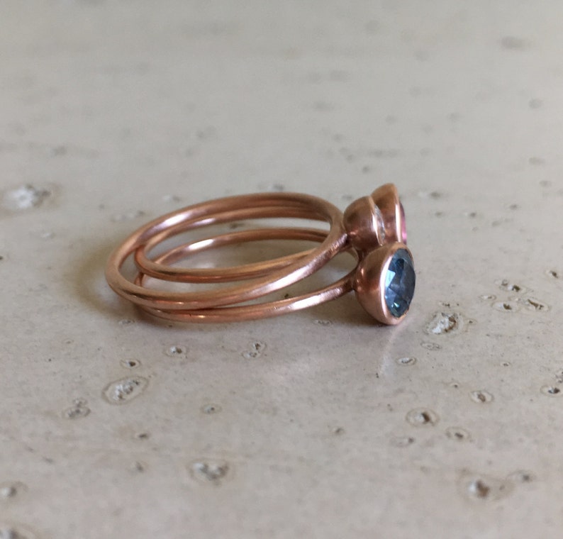 May include: Three copper rings stacked together. Two rings have bezel-set gemstones: one pink and one teal. The rings are on a light-colored, textured surface. The rings are delicate and minimalist.