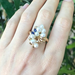 May include: A gold ring with a cluster of white pearls and blue gemstones. The ring is set on a gold band.