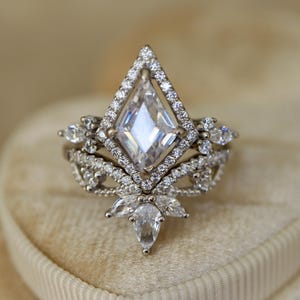 May include: A diamond-shaped engagement ring with a large, clear center stone. The ring is surrounded by smaller diamonds and set in a silver band. The ring has a unique, ornate design with a floral-inspired base.