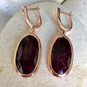 Könnte beinhalten: Ein Paar Ohrringe aus Roségold mit ovalen, dunkelroten Edelsteinen. Die Ohrringe haben ein schlichtes, elegantes Design.