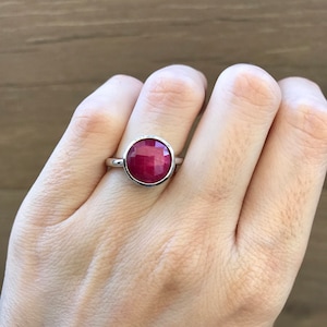 May include: A silver ring featuring a large, faceted, round, ruby-red gemstone. The ring is set in a simple bezel setting, showcasing the vibrant color of the stone. The ring is on a person's finger.