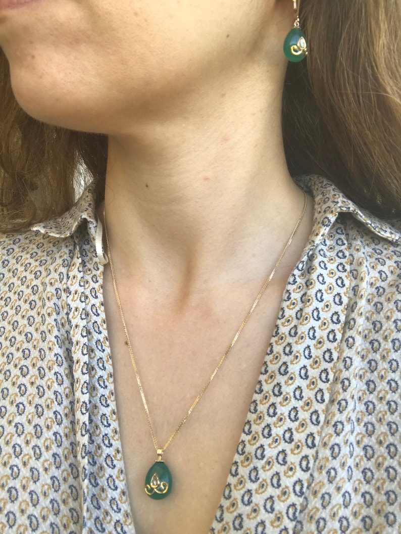 Op de afbeelding: Een gouden ketting met een groene edelsteen hanger. De hanger is druppelvormig en heeft een gouden ontwerp rond de rand. De ketting wordt ook gedragen met bijpassende groene edelsteen oorbellen.