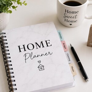 May include: A spiral-bound home planner with a white marble cover featuring the text HOME Planner and a small house icon. The planner includes colored tabs labeled Calendar, Budget, Cleaning, and Meals, accompanied by a black pen and a wooden house sign.