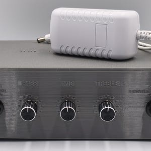 Peut inclure: Un boîtier préamplificateur audio gris rectangulaire avec cinq boutons de commande noirs à centre argenté. Le panneau avant affiche les étiquettes Bypass, Bass, Mid, Treble, Gentle, Subtle et Enhanced. Un adaptateur secteur blanc est posé dessus.