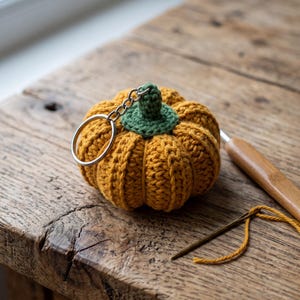 May include: A crocheted pumpkin keychain featuring a vibrant orange body with vertical ribbed texture and a small green stem. A silver metal keyring is attached to the top of the pumpkin, which is positioned next to a wooden crochet hook and a needle.