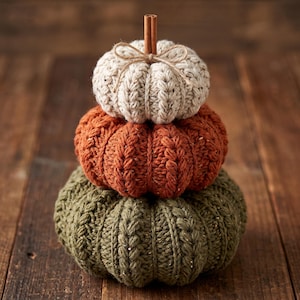 May include: A stack of three crocheted pumpkins in cream, burnt orange, and olive green. Each pumpkin features a ribbed texture and a cinnamon stick stem. The top cream pumpkin is accented with a thin, light brown twine bow tied around the stem.