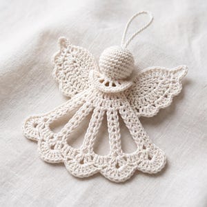May include: A cream-colored crocheted angel ornament featuring a spherical head, open-work wings, and a flared skirt with a scalloped edge. A small crocheted loop is attached to the top of the head for hanging this decorative textile piece.
