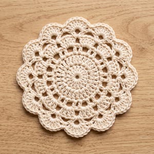 Könnte beinhalten: Ein runder, cremeweißer Häkeluntersetzer mit einem detaillierten, kreisförmigen Muster und einem gewellten, muschelartigen Rand. Das Design strahlt von einem zentralen Punkt aus und zeigt eine Reihe von miteinander verbundenen Schlaufen und Maschen.