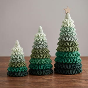May include: Three cone-shaped decorative trees made of crocheted yarn in an ombre color palette ranging from light mint green at the top to dark forest green at the base. The tallest tree is topped with a small, light-colored wooden star.