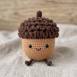 May include: A crocheted acorn toy featuring a textured dark brown cap and a smooth light tan base. The acorn has two small black plastic eyes and a stitched black smile, with two tiny brown crocheted legs extending from the bottom of the base.