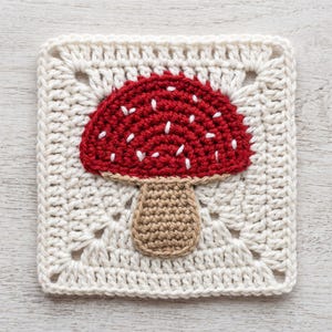 Może przedstawiać: Kwadratowy element szydełkowy typu granny square z centralnym motywem czerwonego grzyba z białymi haftowanymi kropkami na beżowym trzonku. Tło kwadratu wykonane jest z przędzy w kolorze złamanej bieli z klasycznym ażurowym wzorem ściegu.
