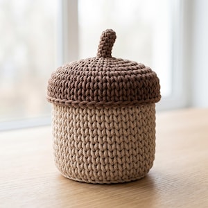 Puede incluir: Un recipiente cilíndrico hecho de hilo de ganchillo con forma de bellota. La base es de color beige claro, mientras que la tapa extraíble es de un marrón más oscuro con un pequeño tallo vertical en el centro. La textura muestra puntos.