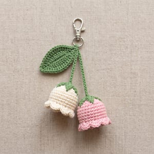 May include: A crochet keyring featuring two bell-shaped flowers in cream and light pink, attached to a green stem with a single green leaf. The flowers hang from a silver-toned metal lobster clasp, showcasing detailed yarn stitching and scalloped edges.
