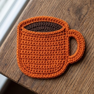 May include: A flat, crocheted coaster shaped like a coffee mug. The main body of the mug is bright orange with a textured stitch pattern, while the top interior section is dark brown, representing coffee. It features a curved orange handle.