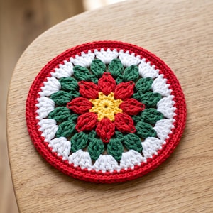 May include: A circular crochet coaster featuring a vibrant floral pattern. The centre is bright yellow, surrounded by layers of red and dark green petals, finished with a white scalloped border and a solid red outer edge made of cotton yarn.