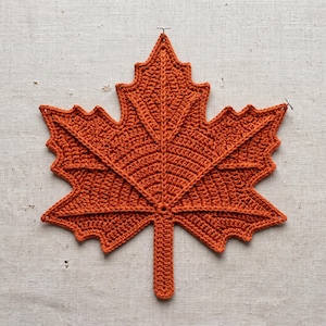 May include: A burnt orange crochet maple leaf with a textured, ribbed surface and a central stem. The leaf features five distinct pointed lobes and intricate stitching that mimics the natural veins of a leaf, creating a flat, decorative shape.