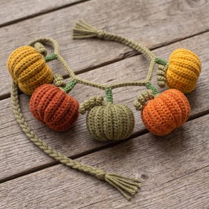 May include: A crochet garland featuring five small pumpkins in shades of yellow, orange, and green. The pumpkins are connected by a braided green cord with small green stems and leaves attached to each, ending in tasseled ends on both sides.