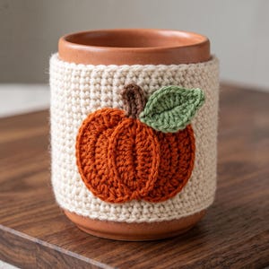 Op de afbeelding: Een terracotta mok omhuld door een crèmekleurige gehaakte hoes. De hoes is voorzien van een oranje pompoenapplicatie met een bruine steel en een groen blaadje, alles vervaardigd met zichtbare garensteken en een textuurrijk, handgemaakt uiterlijk.