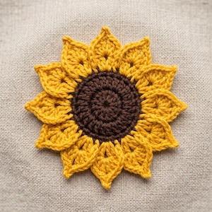 May include: A crocheted sunflower applique featuring a dark brown circular center surrounded by two layers of pointed, bright yellow petals. The flower is crafted with visible yarn stitches, creating a textured, flat, and circular floral design.