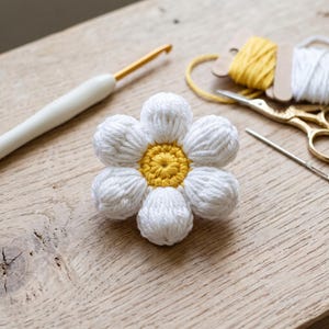 May include: A crocheted flower with six rounded white petals and a textured yellow centre. The flower is accompanied by a white crochet hook, a needle, gold-toned scissors, and spools of yellow and white yarn on a light wood surface.