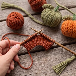 May include: A pair of wooden knitting needles holding a row of rust-colored yarn in a ribbed stitch pattern, accompanied by a ball of matching yarn and a decorative garland featuring crocheted pumpkins in shades of orange, green, and brown.