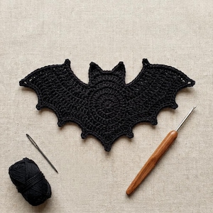 May include: A black crocheted bat applique with pointed ears and scalloped wings. Beside the bat are a small ball of black yarn, a silver metal sewing needle, and a wooden crochet hook with a smooth, tapered handle for crafting projects.