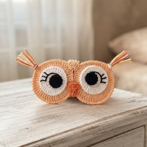 Pode incluir: Uma máscara de dormir de crochê em forma de coruja na cor pêssego, com grandes manchas circulares brancas nos olhos, pupilas pretas e cílios bordados. A máscara inclui um pequeno bico triangular laranja e dois tufos nas orelhas feitos de fios laranja, amarelo e branco.
