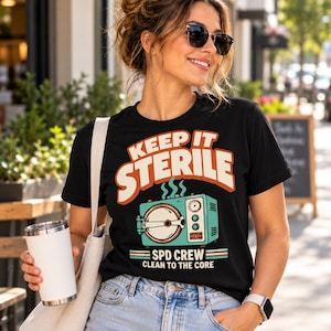 May include: A black short-sleeve t-shirt featuring a graphic of a teal autoclave machine with the text "KEEP IT STERILE" in large orange letters above it and "SPD CREW CLEAN TO THE CORE" in white and teal text below the machine illustration.