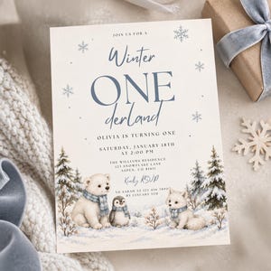 May include: A rectangular invitation card featuring the text Winter ONEderland, Olivia is turning one, and event details. The design includes illustrations of a polar bear, a penguin, and a fox wearing blue scarves in a snowy forest with snowflakes.