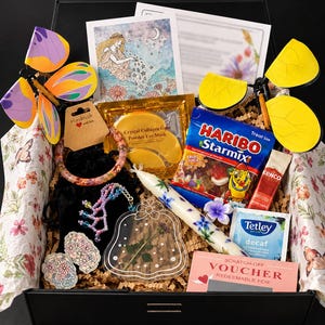 May include: A gift box containing a purple and orange butterfly toy, a yellow butterfly toy, a card with a goddess illustration, gold eye masks, a resin bangle, a beaded chain, a floral candle, Haribo Starmix, Kenco coffee, Tetley tea, and a scratch-off voucher.