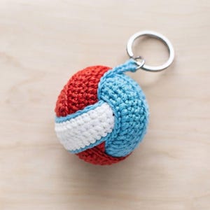May include: A spherical crochet keychain featuring a color-blocked design in red, light blue, and white yarn. The round, textured ball is attached to a circular silver-toned metal split ring, providing a secure way to hold keys or attach to bags.