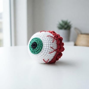 Puede incluir: Un globo ocular de ganchillo esférico con esclerótica blanca, iris verde y pupila negra. La superficie está detallada con costuras de hilo rojo que representan vasos sanguíneos, y el borde posterior tiene un acabado con un borde rojo texturizado.