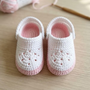 Puede incluir: Un par de zapatos de bebé tejidos a ganchillo a mano con una parte superior blanca con detalles perforados y una suela de color rosa claro. Los zapatos incluyen una puntera redondeada, una tira blanca gruesa en el talón y forro interior.