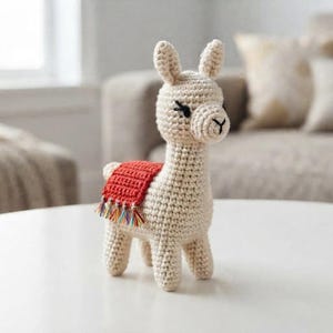 May include: A cream-colored crocheted llama figurine featuring a textured yarn body, upright ears, and a stitched facial expression. The llama wears a small, rectangular red saddle blanket on its back, which is finished with a fringe of multicolored yarn.