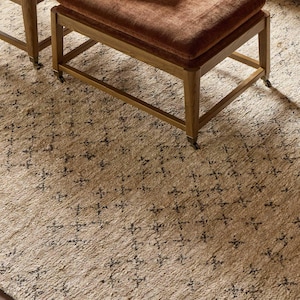 May include: A rectangular ottoman with a rust-colored velvet cushion and a light wood frame on casters. A book titled Japanese Interiors rests on top. The ottoman sits on a beige area rug featuring a repeating dark brown diamond pattern.