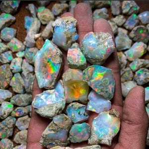 May include: A collection of raw, unpolished opal stones featuring a milky white base with vibrant flashes of iridescent colour, including green, orange, and blue, scattered across the surface of the rough, irregular mineral specimens.