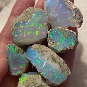 May include: A collection of six raw opal stones featuring a milky white base with vibrant flashes of green, blue, and orange fire. Each stone retains its natural, rough, and unpolished rock matrix edges, showcasing a translucent, iridescent interior.
