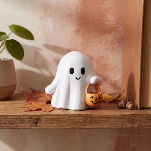 Op de afbeelding: Een wit spookbeeldje met zwarte ogen en een kleine glimlach, dat een miniatuur oranje pompoenemmer vasthoudt. Het spook heeft een gladde, ronde vorm en staat rechtop, met een klassiek lakenachtig uiterlijk voor Halloween.
