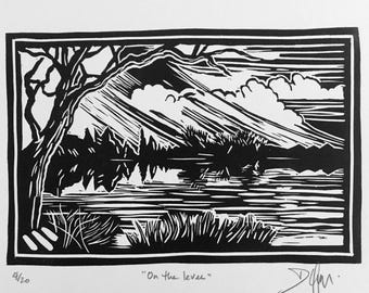 May include: A black and white linocut print featuring a landscape scene with a tree on the left, a body of water, and clouds in the sky. The print is titled "On the levee" with the edition number 4/20 and a signature visible at the bottom.