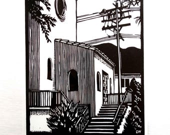 May include: A black and white linocut print depicting a building with arched windows, a circular window, and a staircase leading to an entrance. A utility pole with wires stands nearby, and foliage is visible in the foreground. Signed DY 2014.