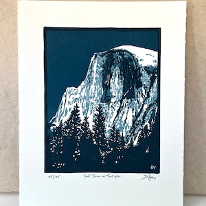 May include: A blue and white linocut print of Half Dome in Yosemite National Park. The print is titled "Half Dome at Twilight" and is numbered 25/25. The artist's signature is in the bottom right corner.