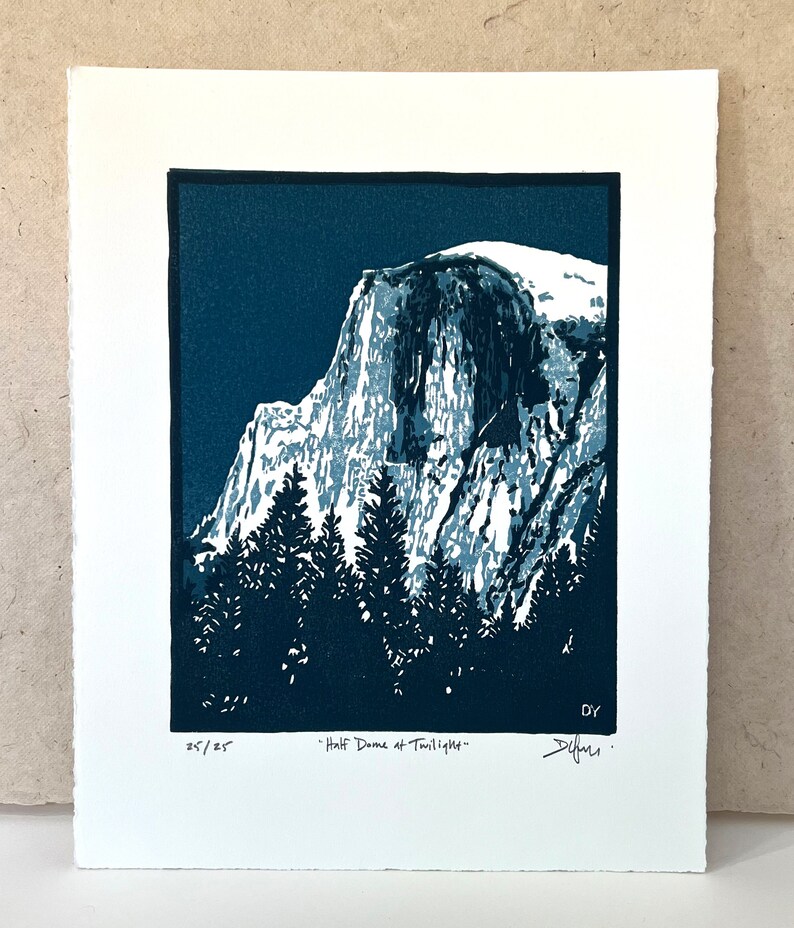 May include: A blue and white linocut print of Half Dome in Yosemite National Park. The print is titled "Half Dome at Twilight" and is numbered 25/25. The artist's signature is in the bottom right corner.