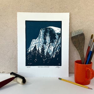 May include: A blue and white linocut print of Half Dome in Yosemite National Park. The print is titled "Half Dome at Twilight" and is signed by the artist.