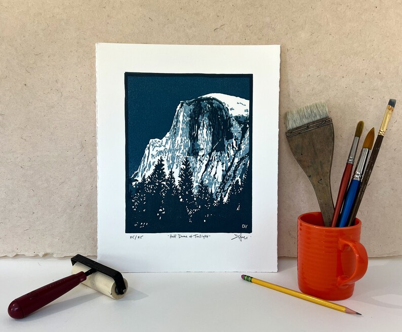 May include: A blue and white linocut print of Half Dome in Yosemite National Park. The print is titled "Half Dome at Twilight" and is signed by the artist.