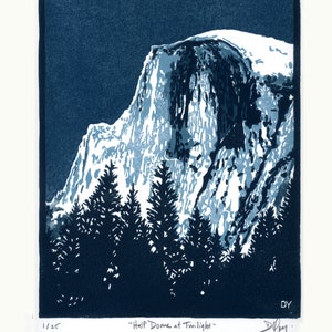 May include: A blue and white linocut print of Half Dome in Yosemite National Park at twilight. The print features a dark blue background with a white snow-capped mountain peak. The text "Half Dome at Twilight" is printed below the image.