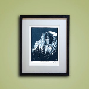 May include: A blue and white linocut print of Half Dome in Yosemite National Park. The print is framed in a black frame and has the title "Half Dome at Twilight" and the artist's signature.