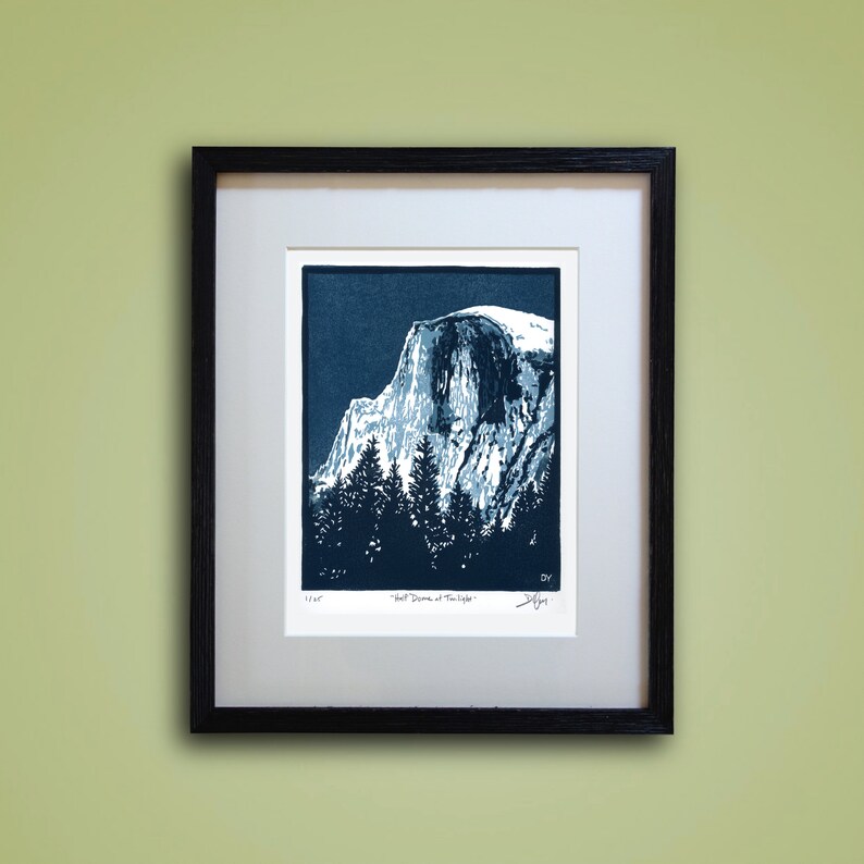 May include: A blue and white linocut print of Half Dome in Yosemite National Park. The print is framed in a black frame and has the title "Half Dome at Twilight" and the artist's signature.