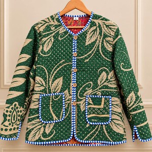 Può includere: Una giacca trapuntata a maniche lunghe verde scuro con un motivo a foglie color marrone chiaro e una trama a piccoli punti bianchi. Presenta un bordo a righe blu e bianche, cinque bottoni in legno e due tasche frontali. La fodera è rossa.