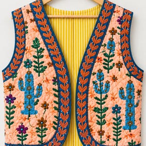Könnte beinhalten: Eine gesteppte ärmellose Weste aus pfirsichfarbenem Stoff mit blauen Kaktus- und bunten Blumenstickereien. Das Kleidungsstück hat einen dunkelblauen Rand mit orangefarbenen Blattmustern und ein gelb-weiß gestreiftes Innenfutter.